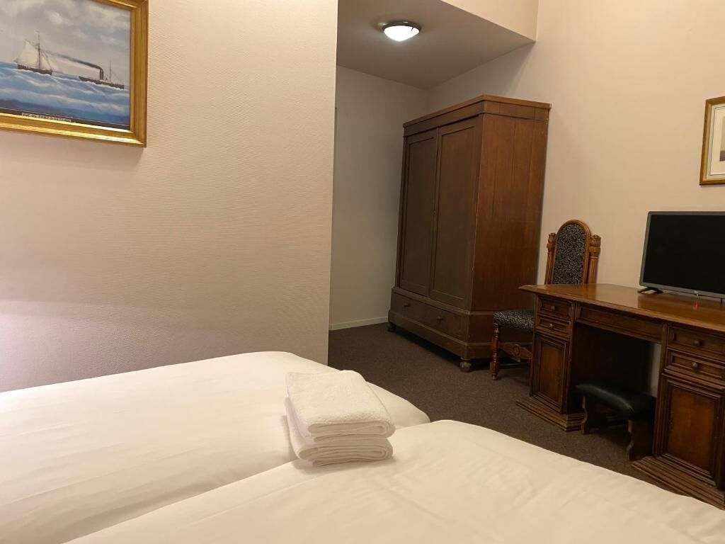 A hotel room with two beds, a wooden wardrobe, a desk with a television, and a painting on the wall.