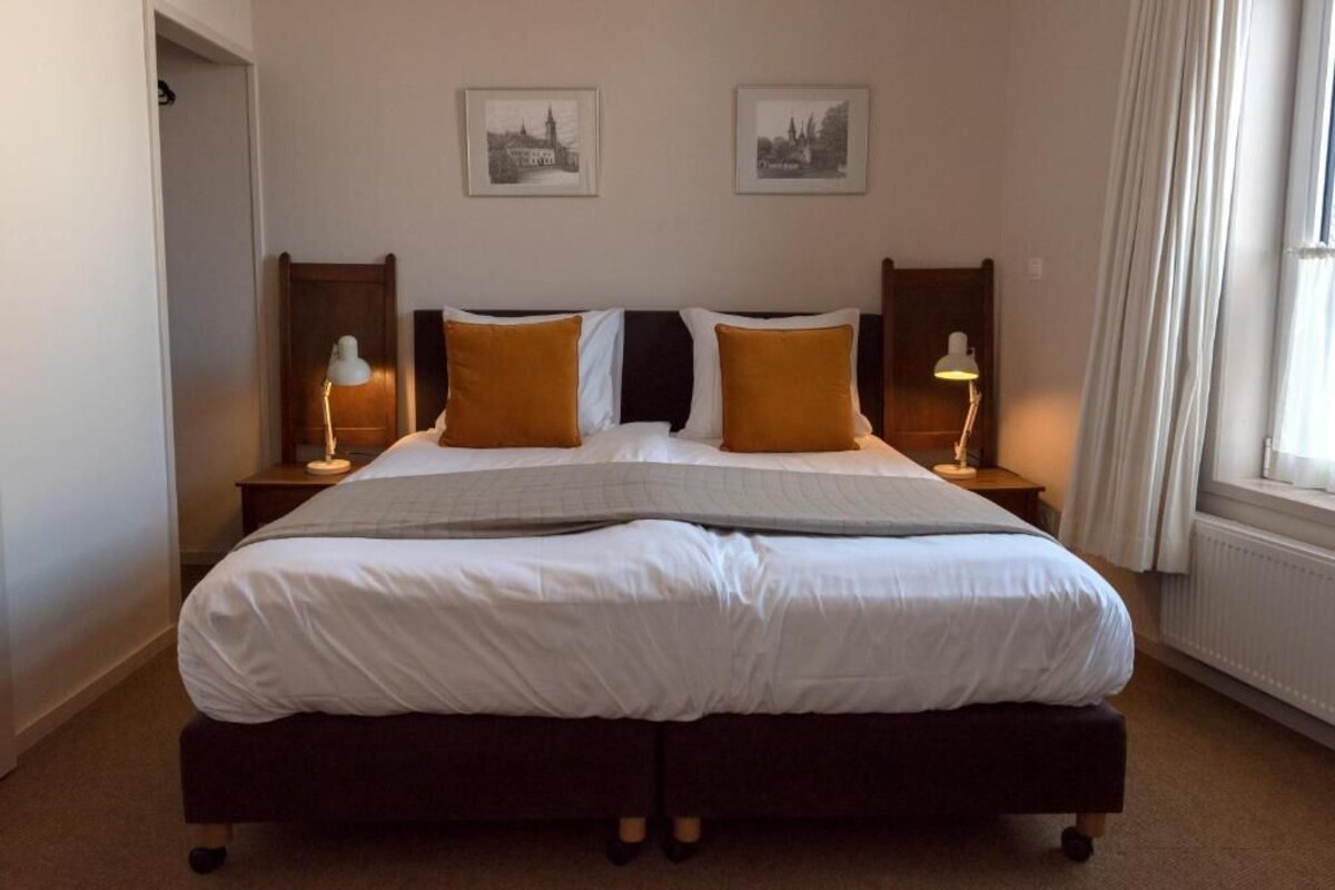 Ein Doppelbett mit weißer Bettwäsche und zwei orangenen Kissen, an beiden Seiten Nachttische mit Lampen, in einem Zimmer mit zwei gerahmten Bildern an der Wand.