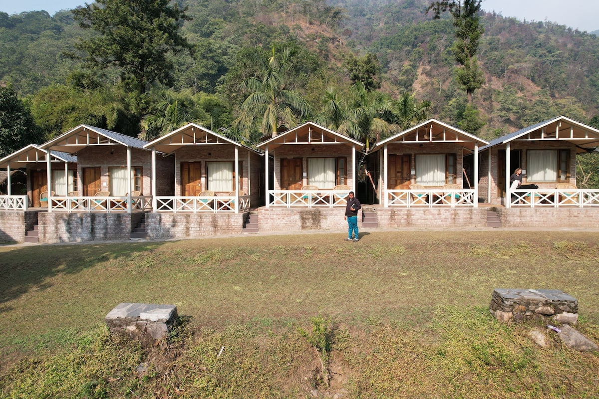 A row of wooden cabins with white railings, set against a backdrop of lush greenery and hills.