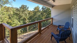 Terraza de madera con ventilador de techo, dos sillas Adirondack azules y vista a un bosque.
