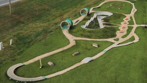 Vista aérea de un parque con senderos sinuosos, estructuras cilíndricas verdes y conos de tráfico naranjas.