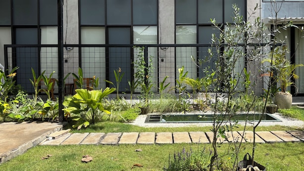 A modern building with large windows, a black metal fence, and a well-maintained garden with various plants and a small water feature. - Olive Athena Guesthouse (Denpasar)