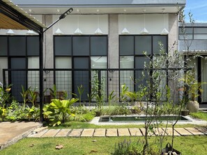 Ein modernes Gebäude mit großen Fenstern, einem schwarzen Metallzaun und einem gepflegten Garten mit diversen Pflanzen und einem kleinen Wasserspiel.