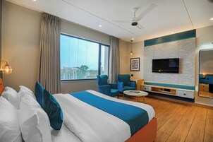 Habitación de hotel moderna con una cama grande, dos sillones azules, una mesita, televisor y vista a la ciudad.