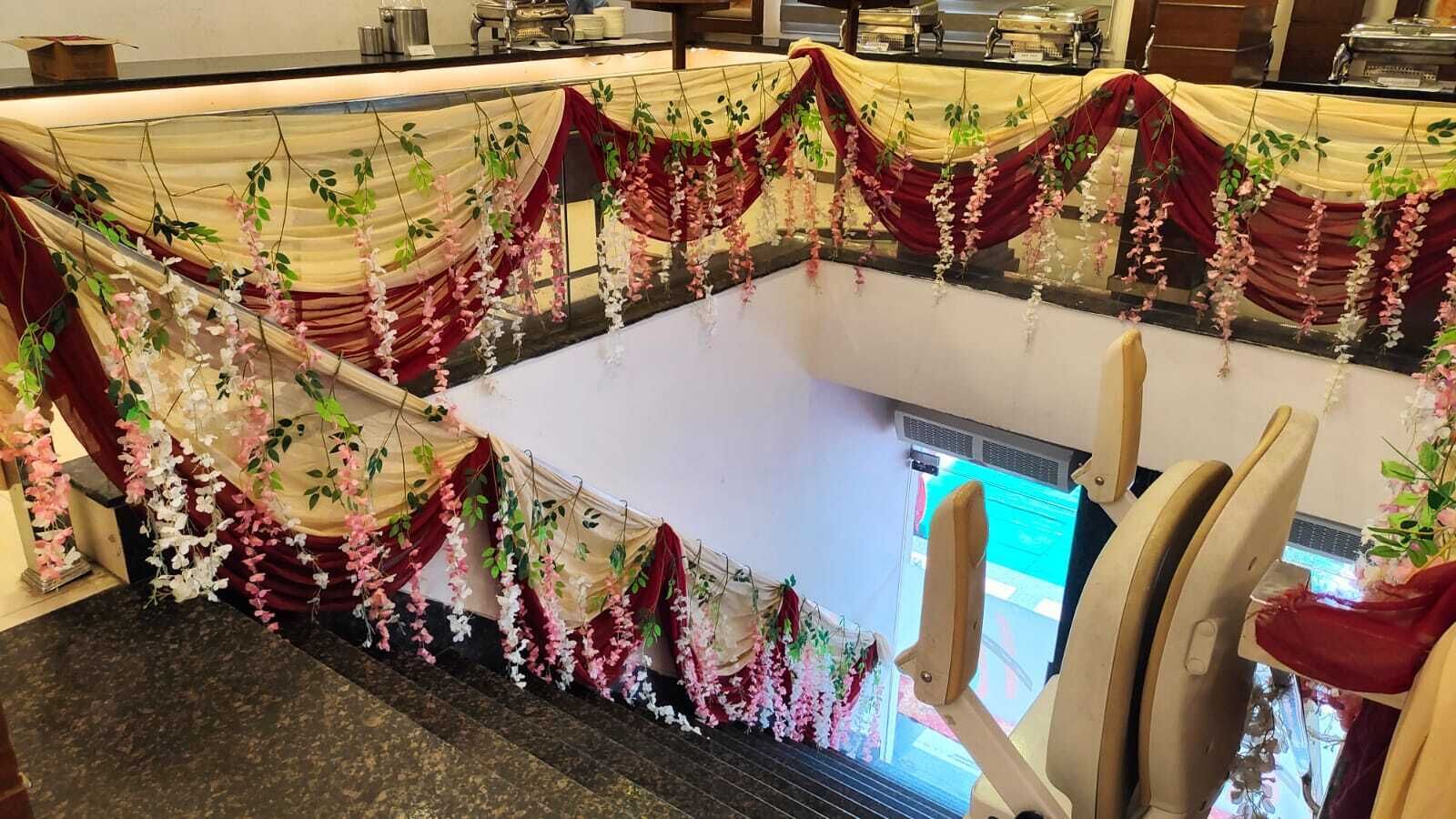 Un vestíbulo de hotel con suelo de mármol, mostrador de recepción y una escalera adornada con cortinas rojas y beige y decoración floral colgante.