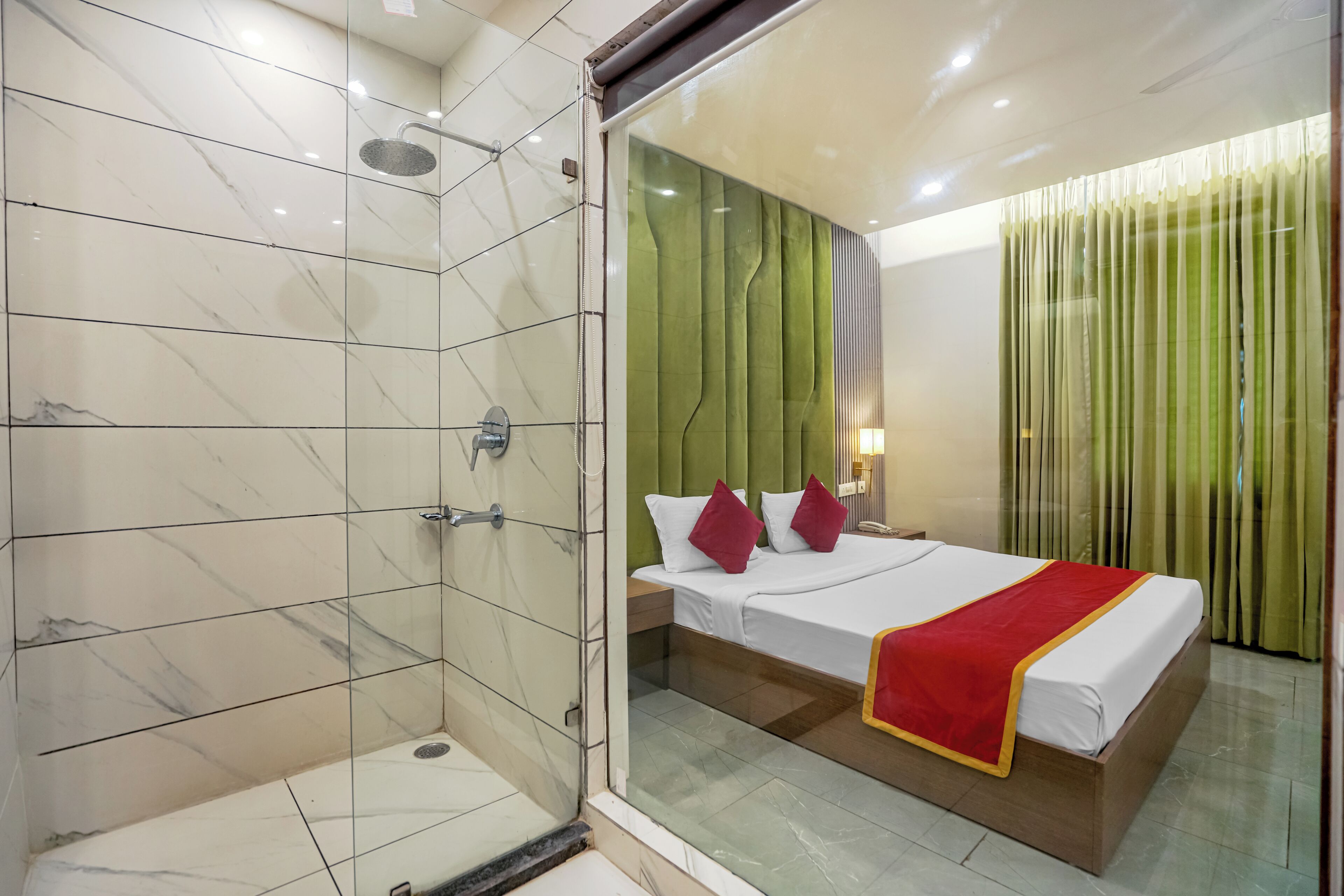 A hotel room with a glass shower enclosure, a bed with white bedding and red accents, and a green curtain.