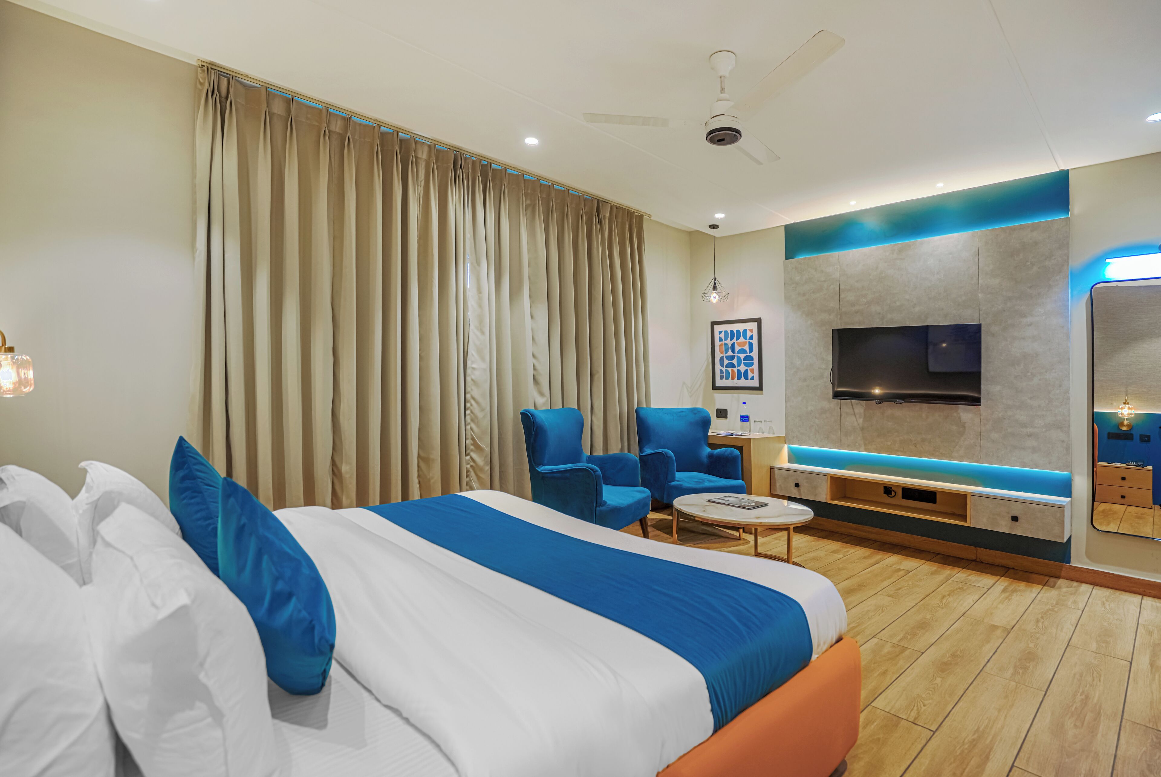 A modern hotel room with a large bed, two armchairs, a small table, a TV, and a minibar.