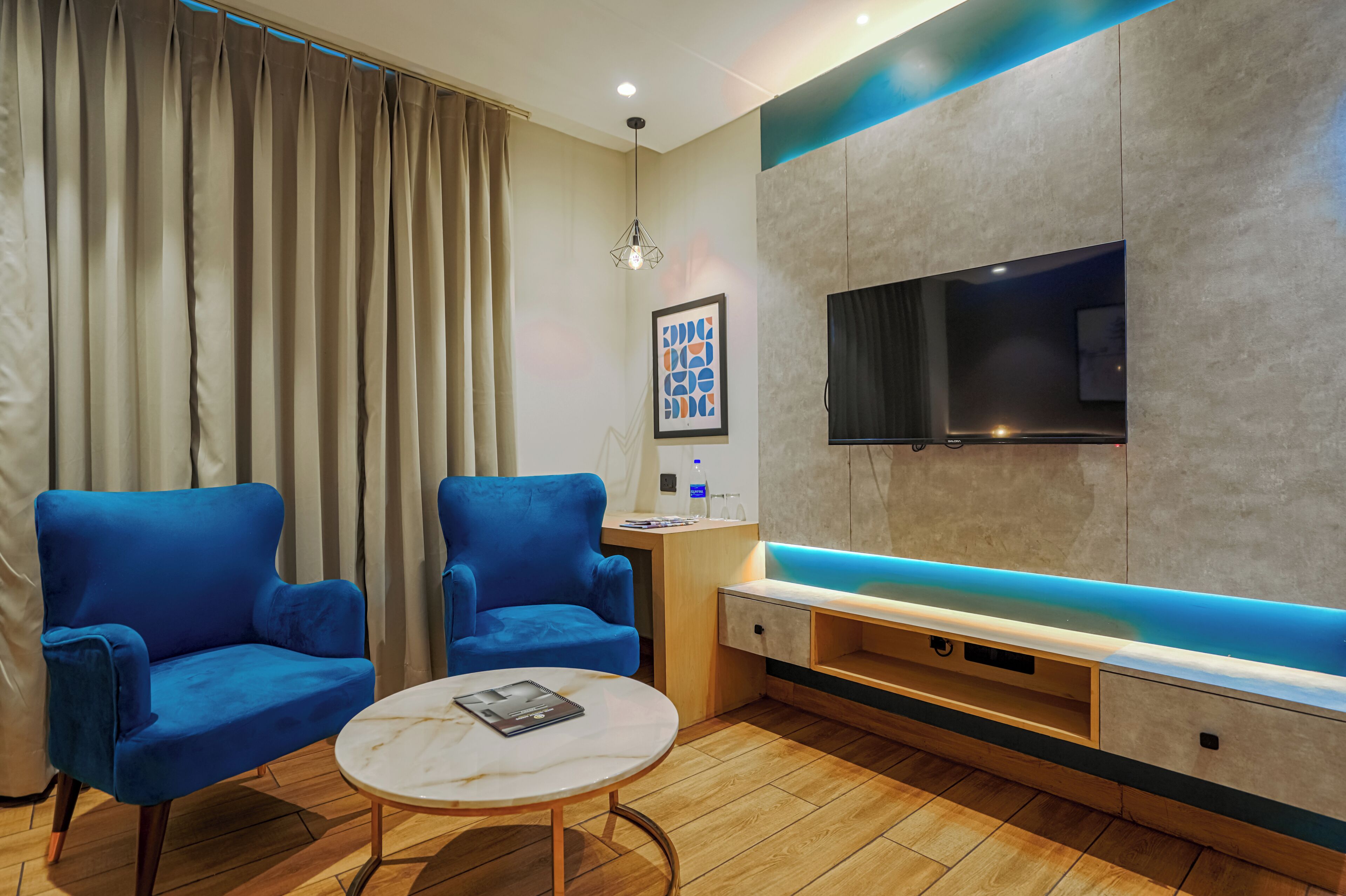 A modern living room with two blue armchairs, a round coffee table, a flat-screen TV mounted on a textured wall, and a blue-lit bench.