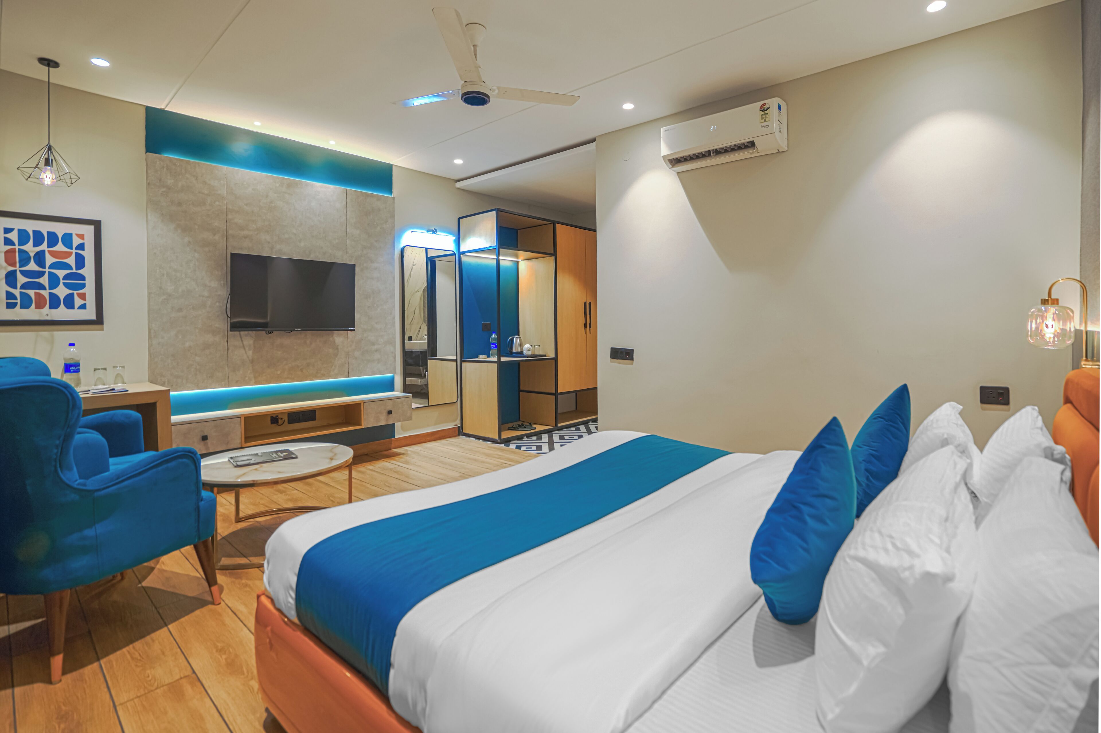 Una habitación de hotel moderna con una cama grande, un área de descanso con una silla azul, un televisor, un espejo y un ventilador de techo.
