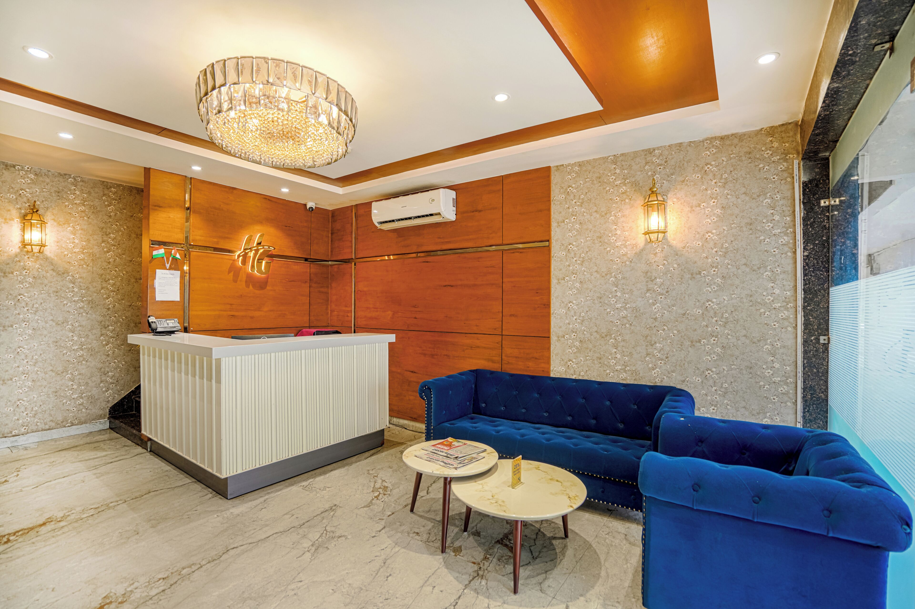 A reception area with a wooden reception desk, a blue sofa, a small round table with a book, and a chandelier.