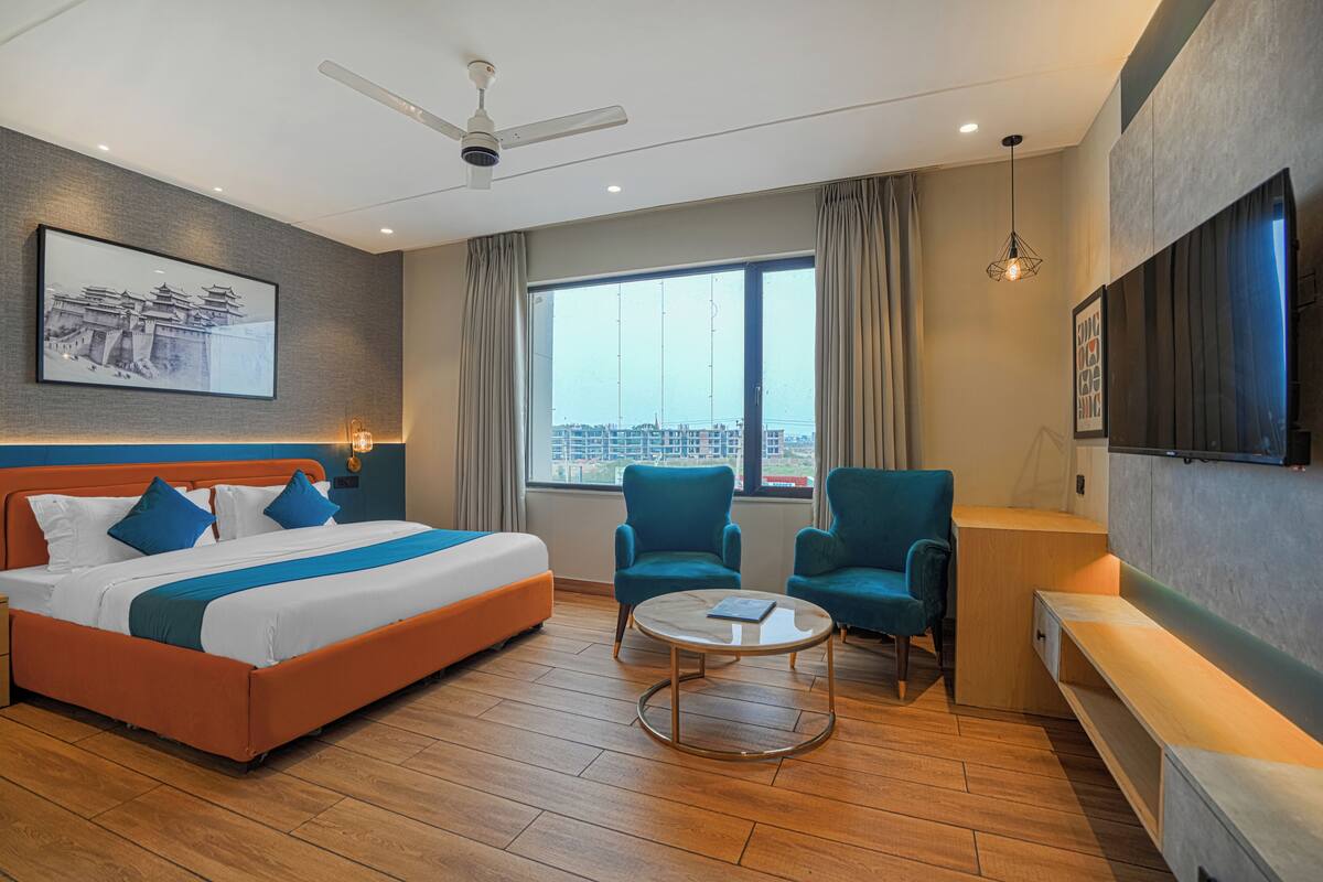 Une chambre d’hôtel moderne dotée d’un grand lit, de deux fauteuils, d’une table basse, d’un ventilateur de plafond, d’une grande fenêtre offrant une vue sur la ville et d’un téléviseur fixé au mur.
