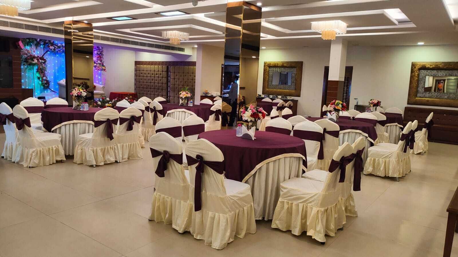 Uma sala de banquetes com mesas redondas cobertas por toalhas bordô, rodeada por cadeiras com capas brancas e fitas escuras.