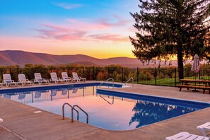 Une piscine entourée d’une clôture et d’arbres, avec des chaises longues et une table de pique-nique, offrant une vue sur des collines lointaines.