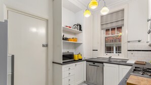 Ett modernt kök med vita skåp, en diskmaskin i rostfritt stål, en bänkskiva i granit och ett fönster med utsikt över en byggnad med tegelstensfasad.