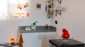 Eine kleine Küche mit Kühlschrank, Spüle und einer kompakten Arbeitsfläche. Ein Fenster mit Vorhang mit Obstmuster, ein Regal mit Küchenutensilien und ein rotes Dekorationsstück auf der Arbeitsfläche.