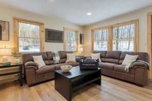 A cozy living room with a brown sectional sofa, a black coffee table with a bowl and a bag, and large windows with a snowy view. - Brookside Mountain Retreat | 13BR | Heated Pool, Hot Tub + Game Room (Stephentown)