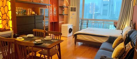 A hotel room with a bed, a dining table with chairs, a kitchen area, and a view of the city through the window.
