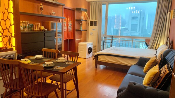 A hotel room with a bed, a dining table with chairs, a kitchen area, and a view of the city through the window. - Beijing CBD - Cozy suite with Queen bed  (Beijing)