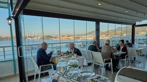 A dining area with tables and chairs, a view of the sea and boats, and a mountainous landscape in the background. - MİA HİLL SUİTES OTEL (Muğla)