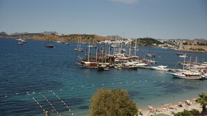 A marina with multiple boats docked, a beach with sun loungers, and a hill with buildings in the background. - MİA HİLL SUİTES OTEL (Muğla)