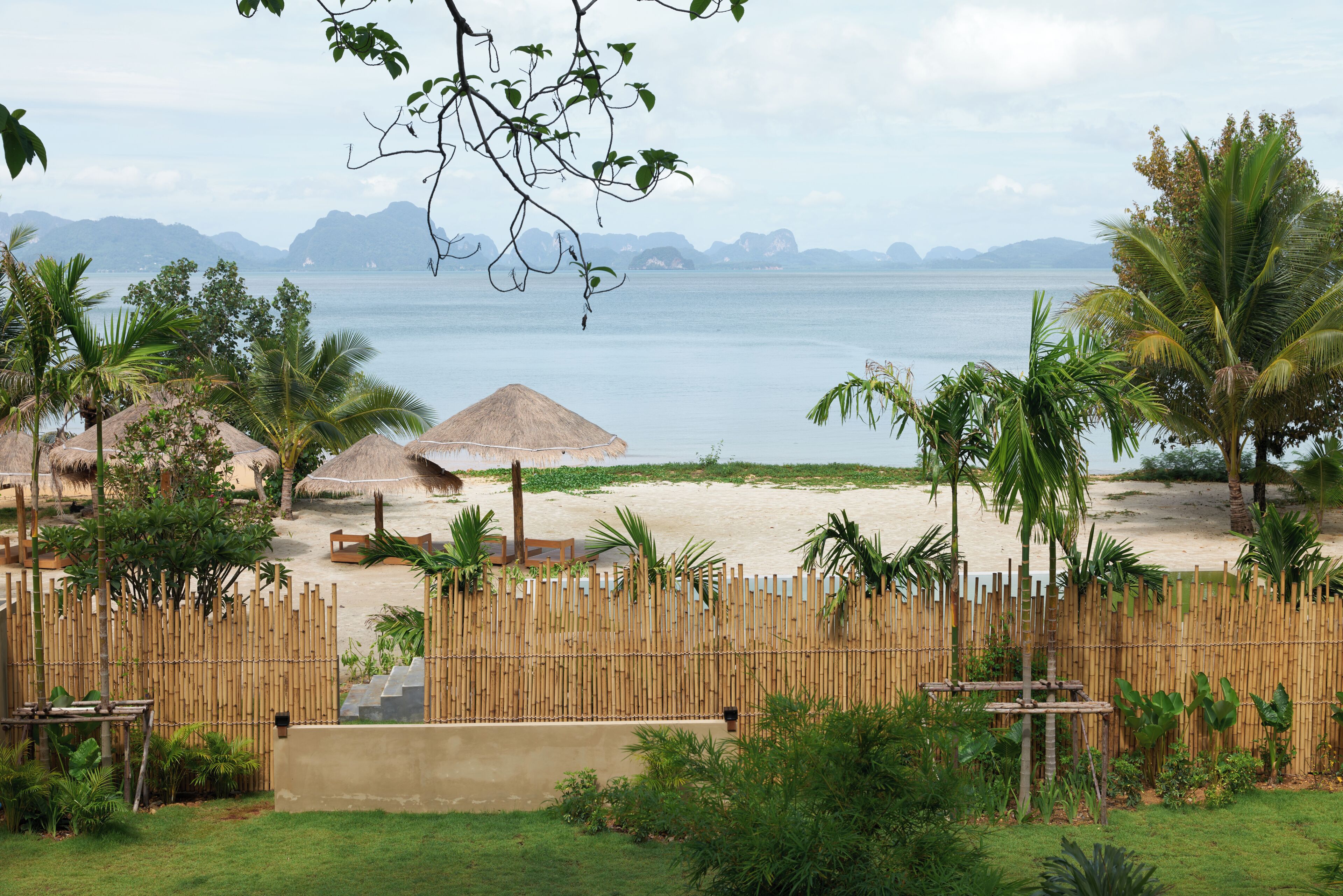 A beach view with a bamboo fence, palm trees, and thatched-roof huts.