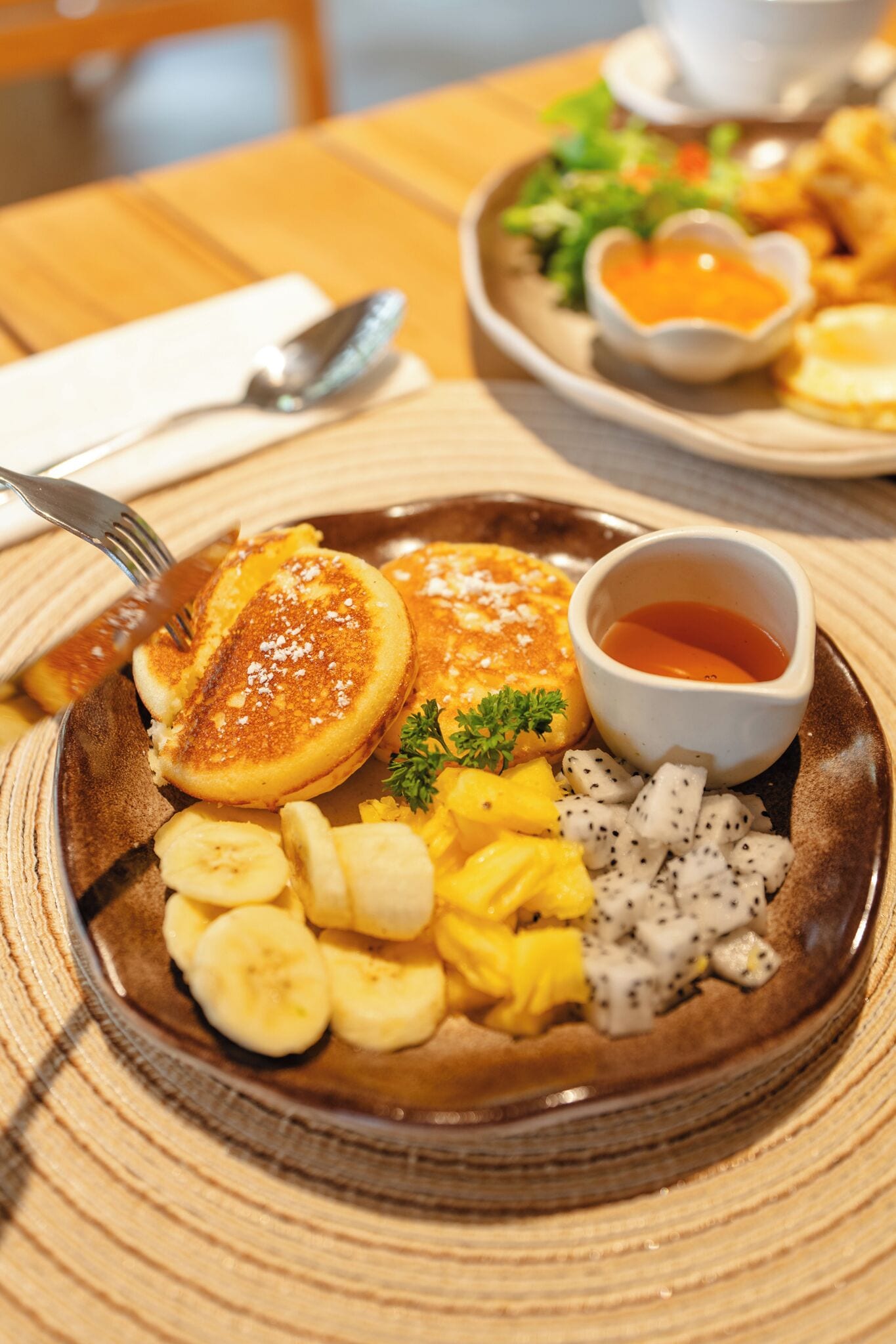 Un plato de desayuno con panqueques, plátanos, piña y un tazón pequeño de almíbar.
