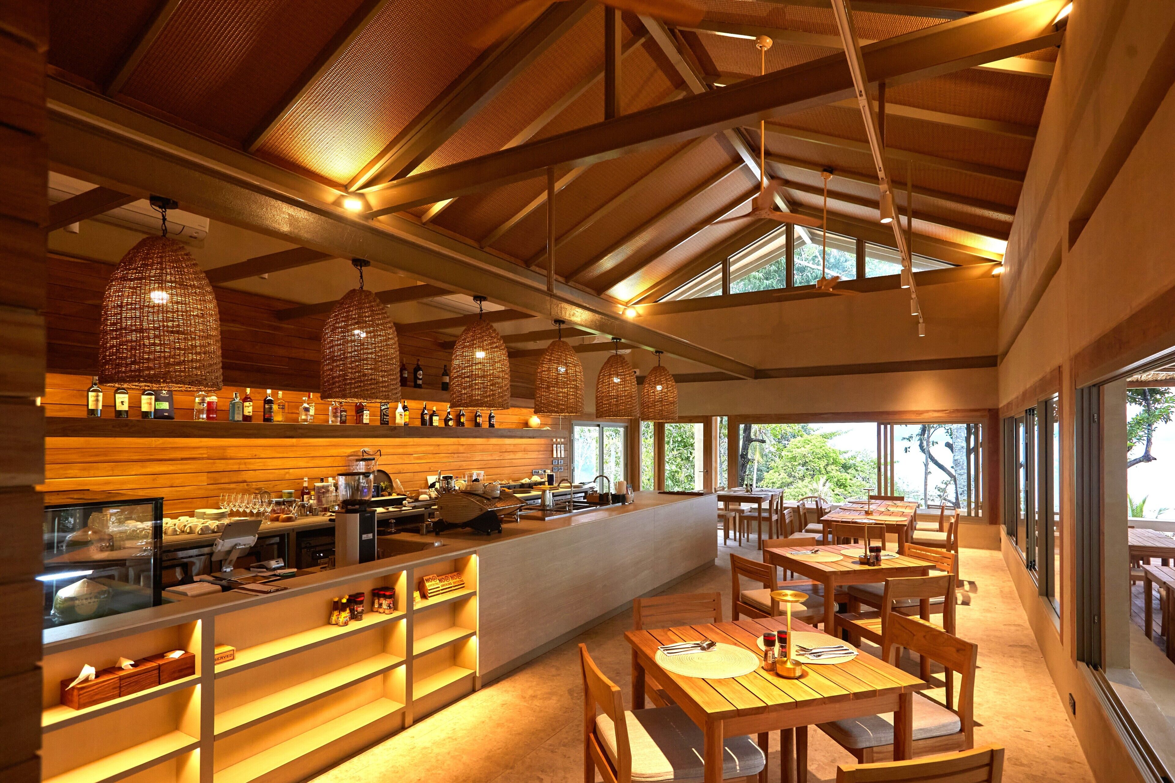 Un restaurant au décor intérieur en bois, agrémenté de lustres en osier suspendus, doté d’un bar et de grandes fenêtres laissant entrer la lumière naturelle.