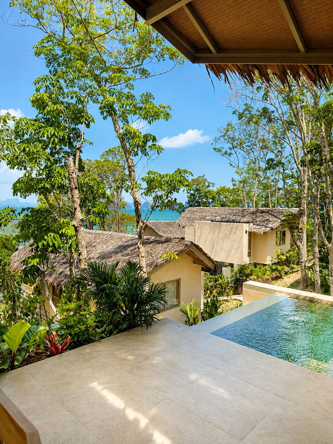 A pool area with a view of the ocean, surrounded by tropical vegetation and thatched-roof buildings.