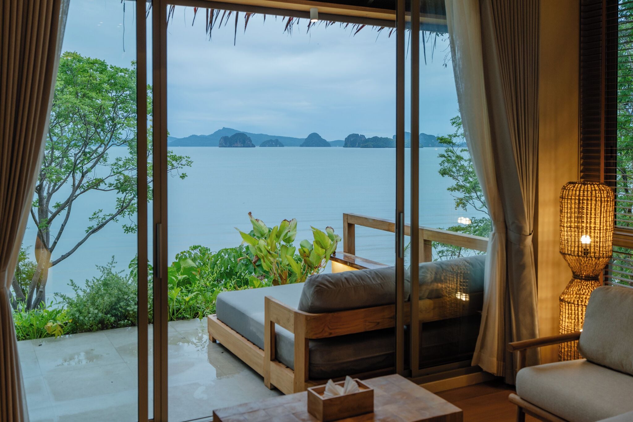 A room with a view of a body of water, a wooden bench, a table, and a lamp.
