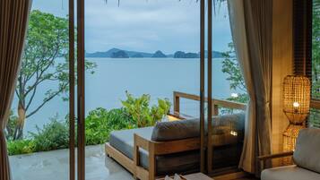A room with a view of a body of water, a wooden bench, a table, and a lamp.