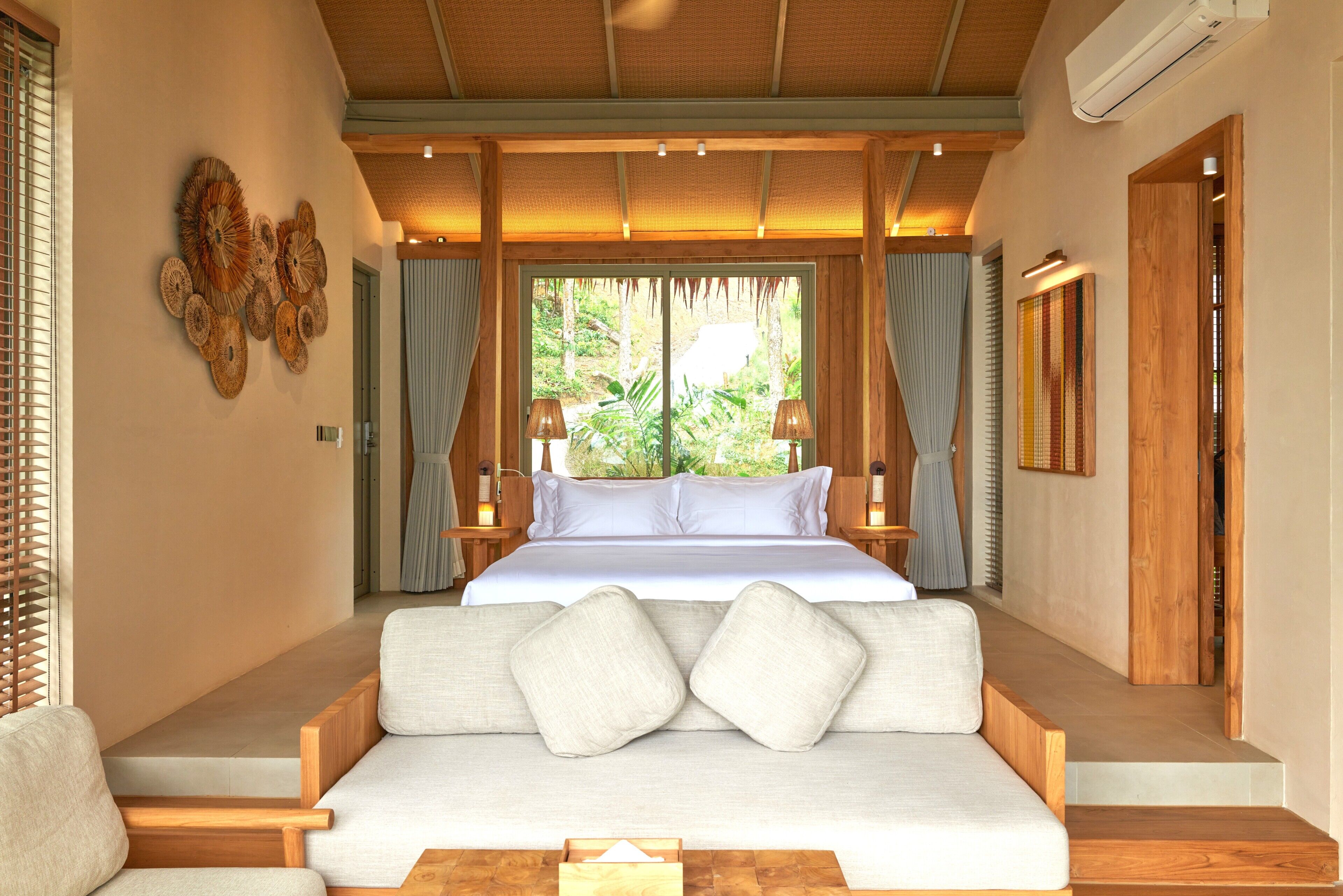 A bedroom with a bed, a sofa, a wooden bench, a wall art piece, and a window with curtains.