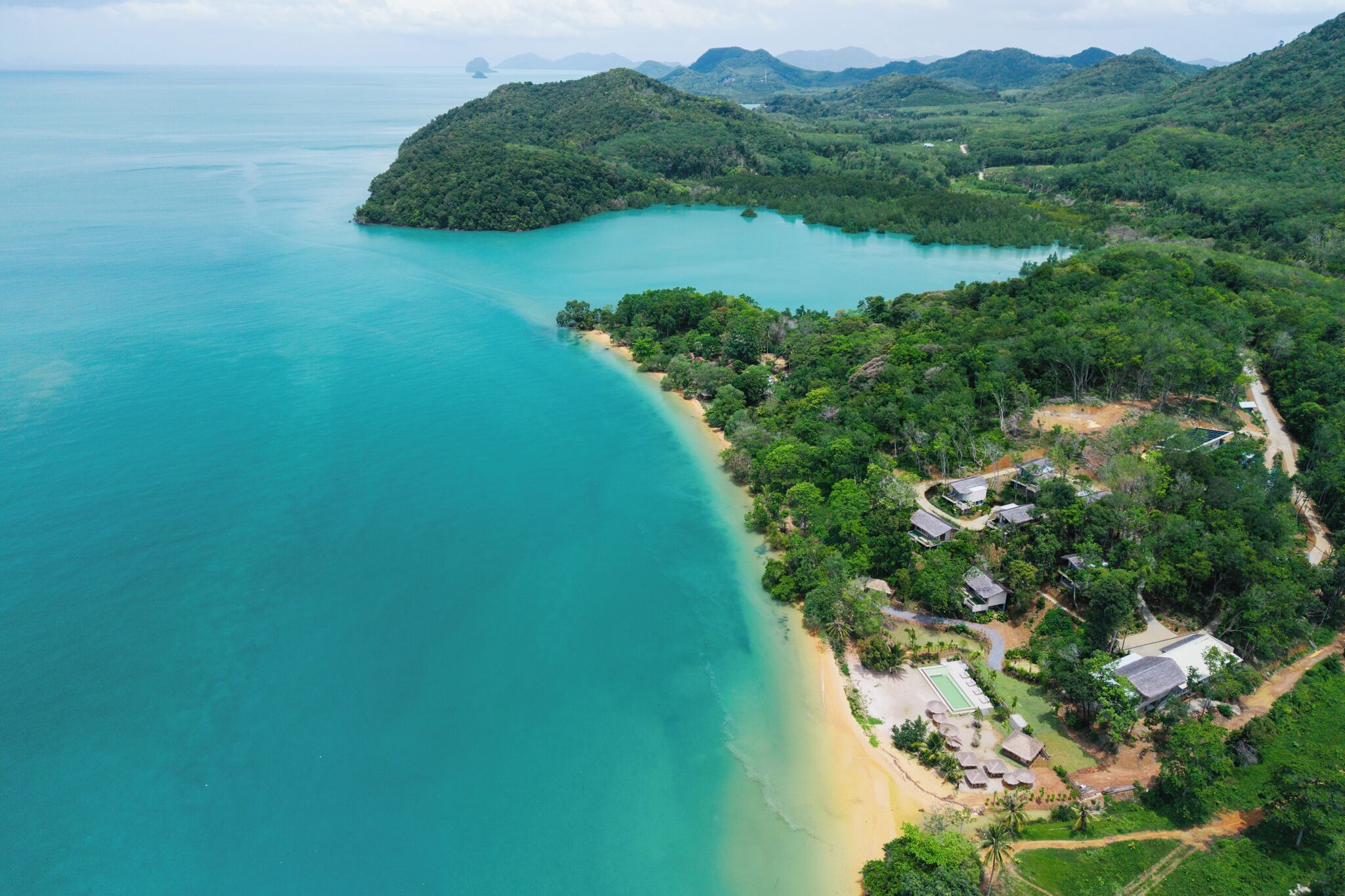 An aerial view of a coastal area with a beach, lush greenery, and a small settlement with buildings and pathways.