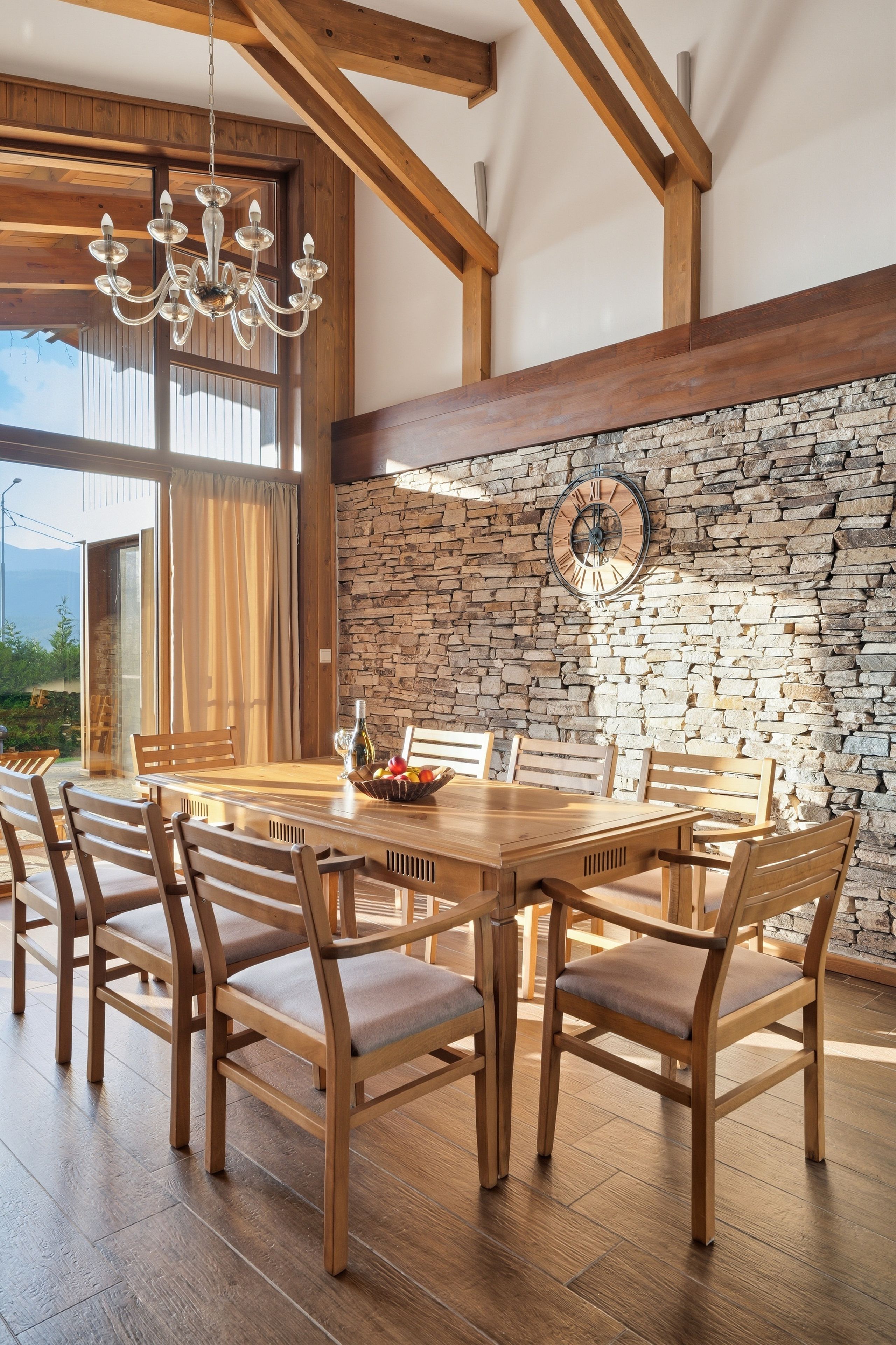 A dining area with a wooden table and chairs, a stone wall with a clock, and a chandelier.