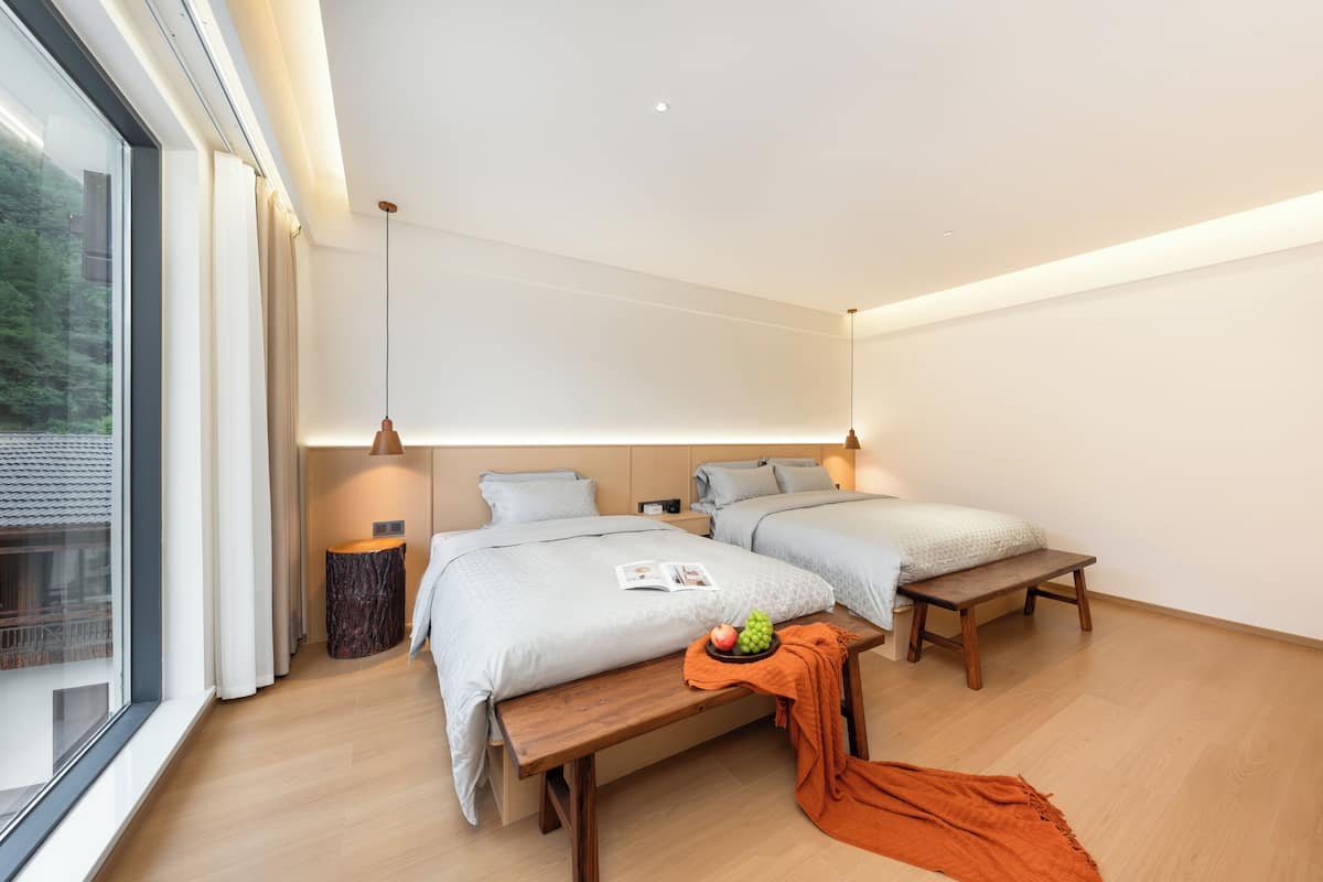 Ein modernes Hotelzimmer mit zwei Betten, einer Holzbank, einem Tisch mit Obst und Blick auf Grün hinter dem Fenster.