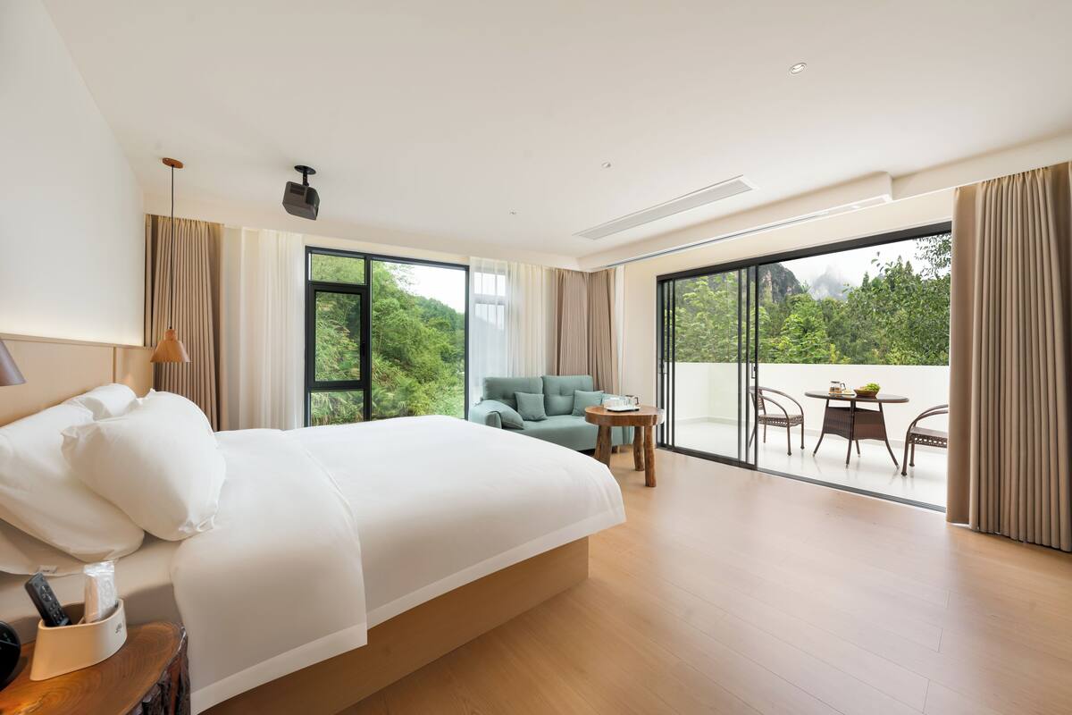 Ein Hotelzimmer mit einem großen Bett, einer Sitzecke mit Tisch und einem Balkon mit Blick auf Grünflächen.