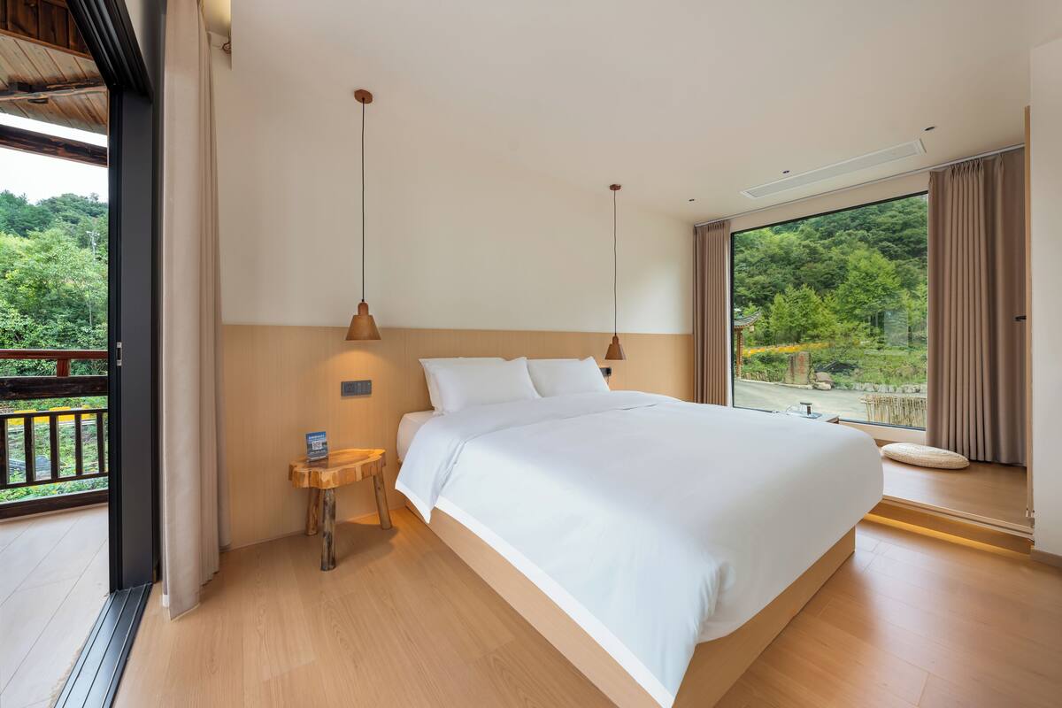 Ein Schlafzimmer mit einem großen Bett, Holzboden, einem kleinen Nachttisch und Blick auf Grün hinter einem großen Fenster.