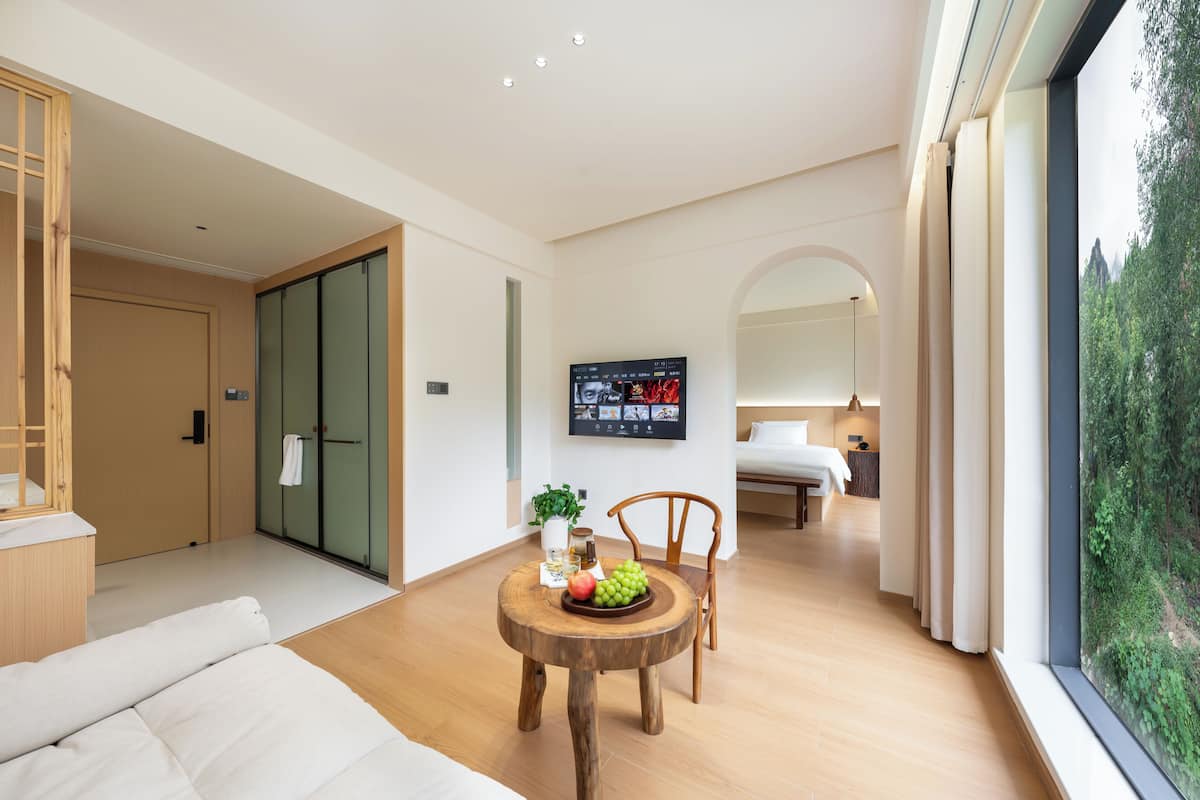 Ein modernes Hotelzimmer mit Holzboden, einem Esstisch mit Obst, einer Couch, einem Fernseher und einem großen Fenster mit Blick auf Grünflächen.