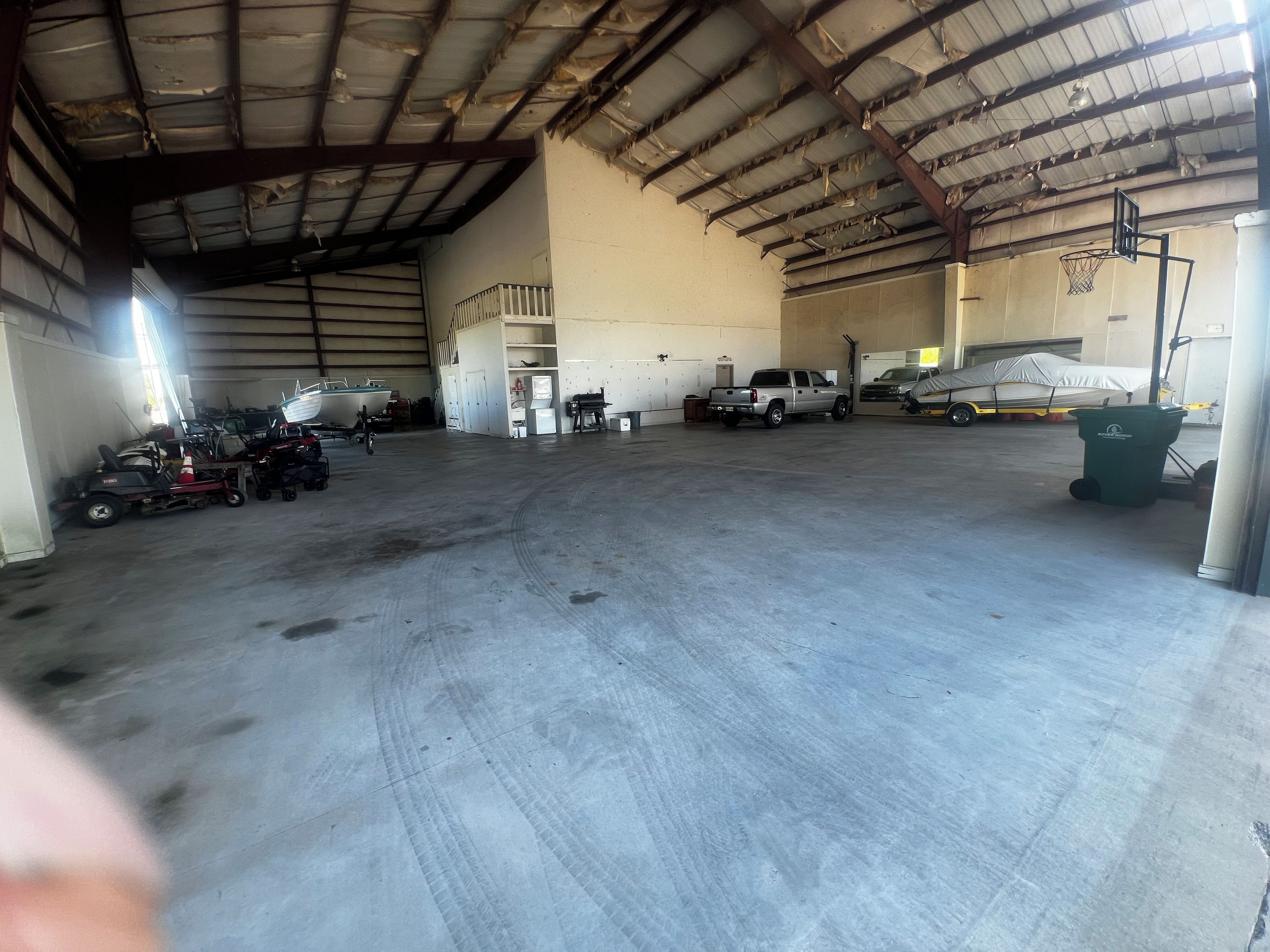 A large indoor space with a high ceiling, concrete floor, and various items including a truck, a covered vehicle, and a green trash bin.