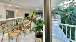 Une cuisine moderne avec des meubles blancs, un réfrigérateur en acier inoxydable et un coin repas doté de chaises en osier et d’une table en verre.