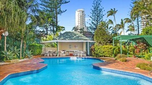 Une piscine dotée de chaises longues et d’un espace salon couvert, entourée d’une végétation tropicale et avec un grand immeuble en arrière-plan.