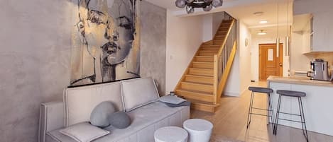 A modern living room with a grey sofa, a large abstract painting, and a staircase leading to a kitchen area.