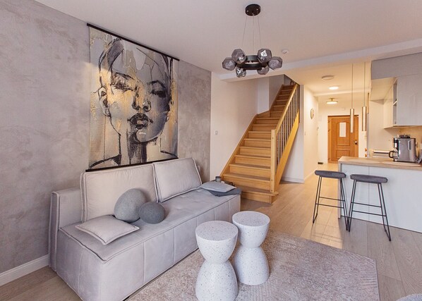 A modern living room with a grey sofa, a large abstract painting, and a staircase leading to a kitchen area.
