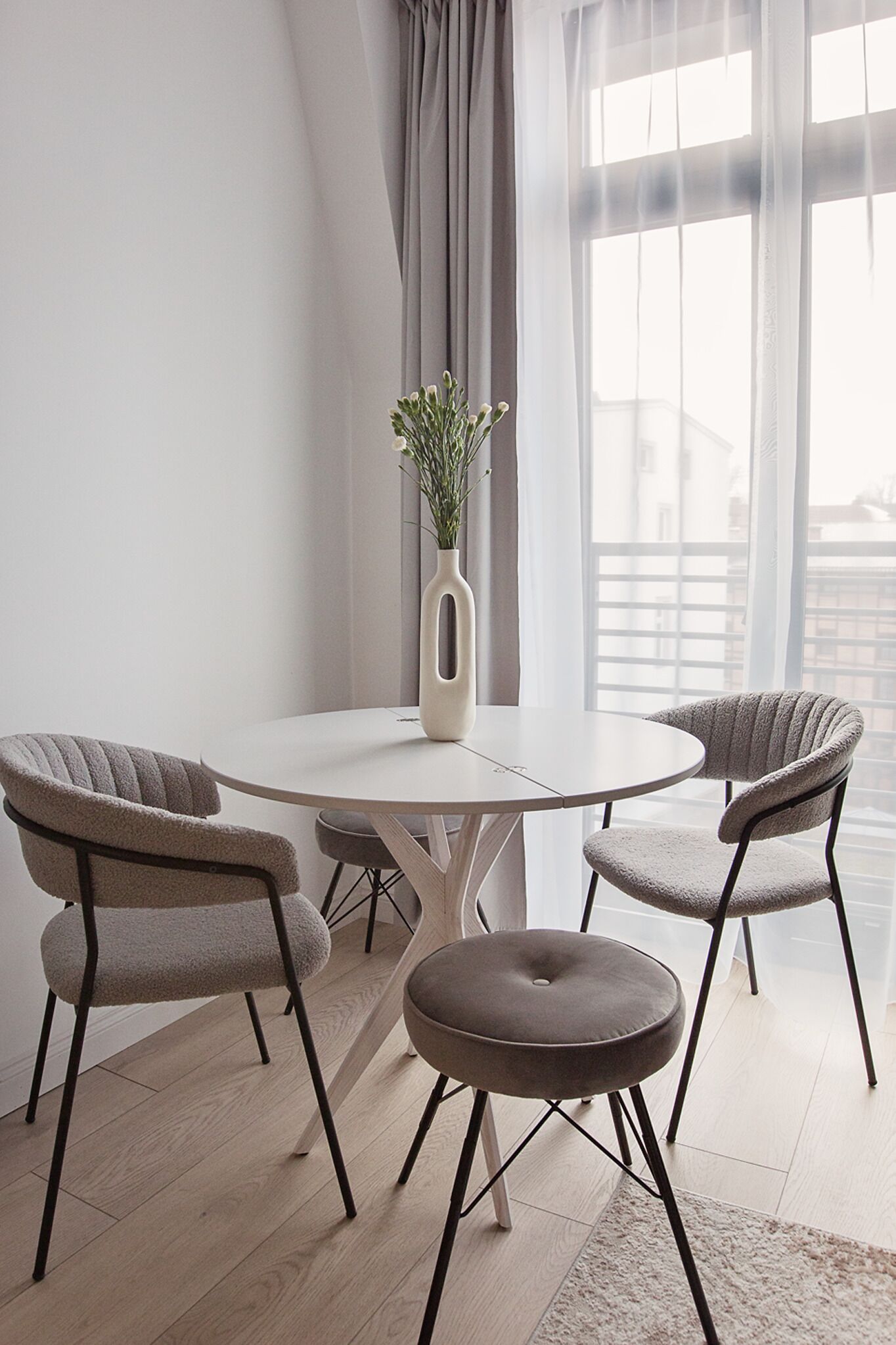 Un comedor moderno con una mesa redonda y cuatro sillas, un florero con plantas verdes, y vistas a través de amplios ventanales.