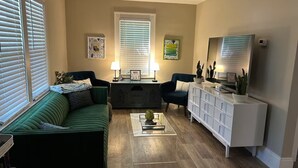 A living room with a green sofa, two armchairs, a coffee table, and a white cabinet. There are two windows with blinds, a lamp, and a TV on a stand. - Stylish and comfortable! House close to downtown Akron. (Akron)