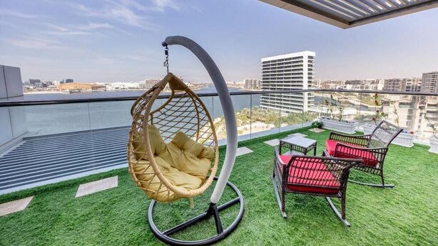 Un balcon agrémenté d’un revêtement de sol en gazon synthétique, d’un fauteuil suspendu et de deux fauteuils en osier avec des coussins rouges.