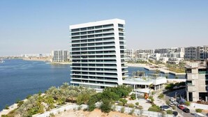 Un hôtel moderne de grande hauteur, doté de nombreux balcons, situé près d’un front de mer bordé de palmiers et d’une plage de sable.