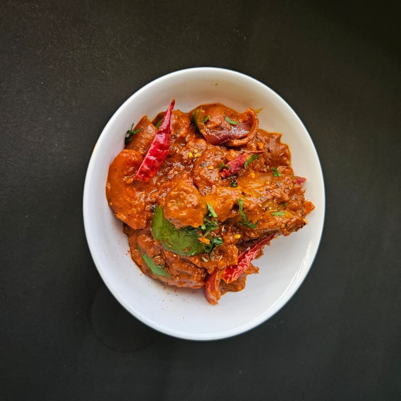 A bowl of curry with visible red chilies and green herbs on a dark surface.