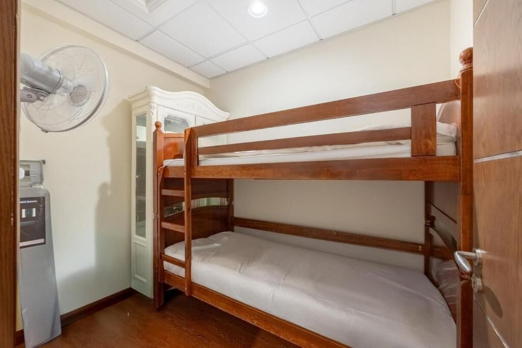 A bunk bed room with a wooden frame, a white mattress, a wooden wardrobe, and a wall-mounted fan.