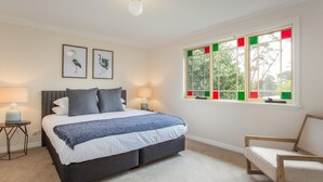 A bedroom with a bed, bedside tables, a chair, a window with colored panes, and wall art. - Stunning Katoomba Home with Lush Garden&Playground (Katoomba)