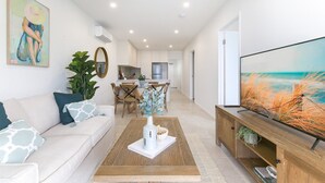 Un salon moderne avec un canapé, une table basse et un téléviseur. On aperçoit à l’arrière-plan un coin repas avec une table et des chaises.
